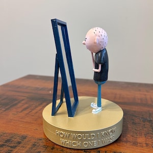 May include: A 3D-printed figurine of a bald person looking into a blue framed mirror. The figure is wearing a black shirt and blue pants, standing on a tan base with the text "HOW WOULD I KNOW WHICH ONE I WAS?"