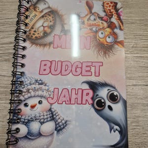 Könnte beinhalten: Ein spiralgebundenes Notizbuch mit einem farbenfrohen Cover, das Cartoon-Illustrationen zeigt. Das Cover zeigt einen Leoparden mit Weihnachtsmütze, einen Schneemann, eine Giraffe und die Worte "Mein Budget Jahr" in Rosa. Das Notizbuch liegt auf einer Holzoberfläche.