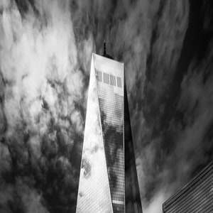 Puede incluir: Fotograf&iacute;a en blanco y negro de un rascacielos alto con un dise&ntilde;o angular distintivo. La fachada de cristal refleja el cielo, que est&aacute; lleno de nubes oscuras y arremolinadas.