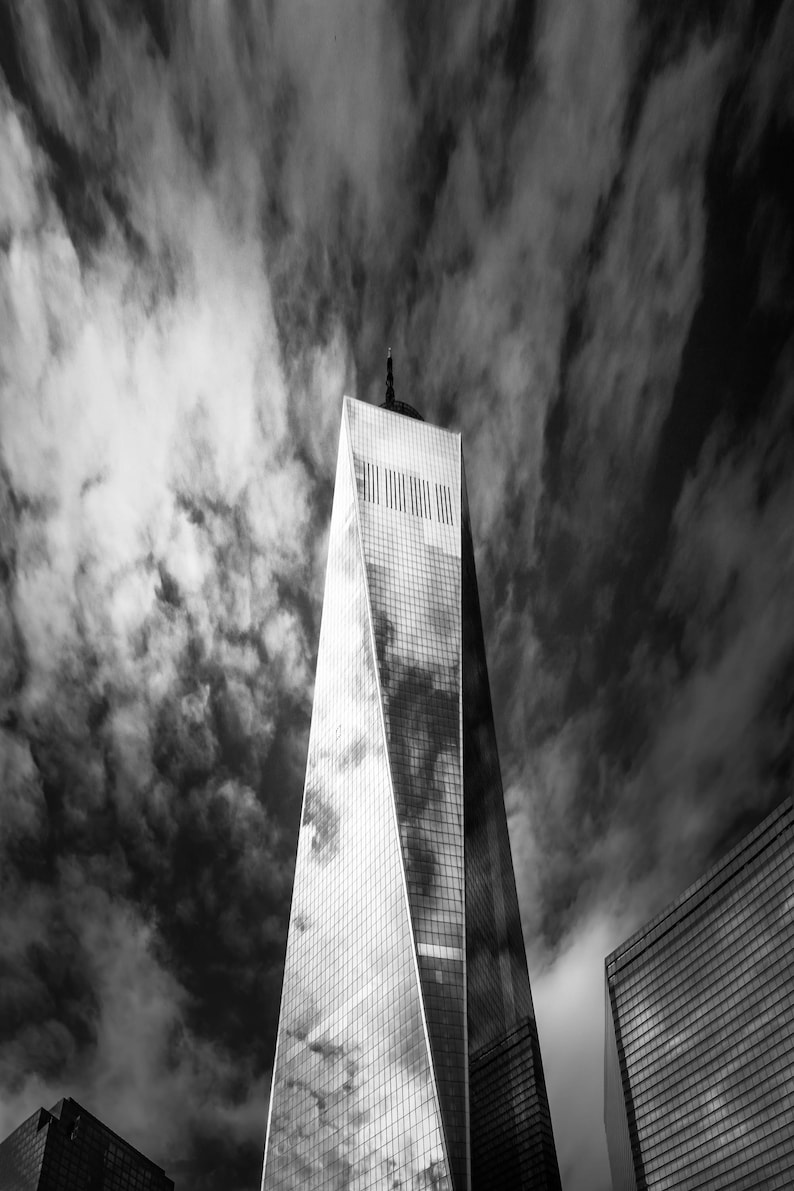 Puede incluir: Fotograf&iacute;a en blanco y negro de un rascacielos alto con un dise&ntilde;o angular distintivo. La fachada de cristal refleja el cielo, que est&aacute; lleno de nubes oscuras y arremolinadas.