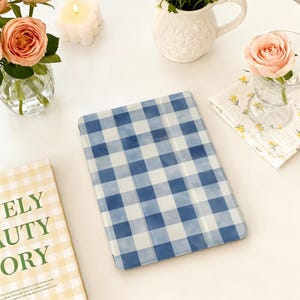 Op de afbeelding: Een tablet hoesje met een blauw en wit ruitpatroon. De hoes ligt op een wit oppervlak, omringd door bloemstukken, kaarsen en een boek met de tekst "ELY BEAUTY STORY".