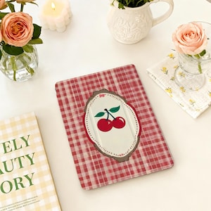 May include: A red and white checkered tablet case with a cherry design. The case features a white oval with a pair of red cherries and green leaves. The background is a red and white gingham pattern.