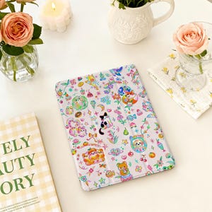 Puede incluir: Una funda blanca para tableta con un patrón colorido y caprichoso de animales de dibujos animados, flores y alimentos. La funda presenta un gato blanco y negro, un zorro y un oso. El diseño incluye estrellas, corazones y lazos.