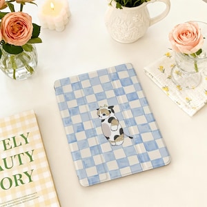 Puede incluir: Una funda para tableta con un patrón de cuadros azules y blancos y una ilustración de gato de dibujos animados. La funda está sobre una superficie blanca, rodeada de flores y una vela.