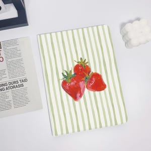 Könnte beinhalten: Ein Notizbuch mit einem grün-weiß gestreiften Muster und einer Aquarellillustration von drei roten Erdbeeren. Die Erdbeeren haben grüne Blätter und sind auf dem Einband des Notizbuchs angeordnet.