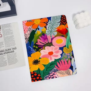 Puede incluir: Una cubierta de cuaderno colorida con un patrón floral. El diseño presenta flores y hojas rosas, amarillas, rojas, azules, verdes y negras sobre un fondo azul oscuro.
