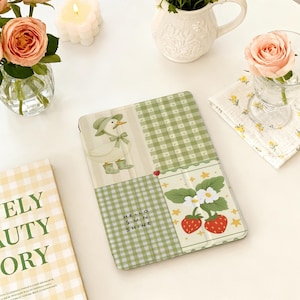 Puede incluir: Una funda para tableta con estampado de cuadros verdes y blancos, con un pato y fresas. El texto "Hello Sun Shine" está presente. Flores y una vela en el fondo.