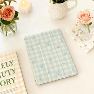 Peut inclure: Une housse de tablette vert clair avec un motif vichy et de petites illustrations de fleurs violettes. L'étui est ouvert, révélant l'intérieur. L'image est mise en scène avec des fleurs, des bougies et un livre.