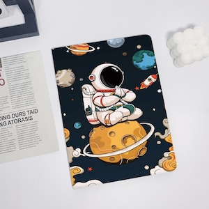 May include: An astronaut in a white spacesuit sits in a meditative pose on a yellow moon with a ring around it. The astronaut is surrounded by stars, planets, and a rocket ship. The background is a dark blue.