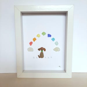 Puede incluir: Una obra de arte enmarcada que presenta un arcoíris hecho de vidrio de mar de colores, con una figurita de perro marrón sentada debajo. El perro está mirando hacia arriba al arcoíris, y hay dos nubes blancas a cada lado del perro. La obra de arte está firmada en la esquina inferior derecha.