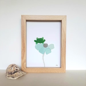 Peut inclure: Une œuvre encadrée représentant une grenouille verte perchée sur une fleur bleu clair. La fleur est faite de verre de mer, avec un centre gris et une fine tige noire. L'œuvre est dans un cadre en bois brun clair. Une coquille d'huître est au premier plan.