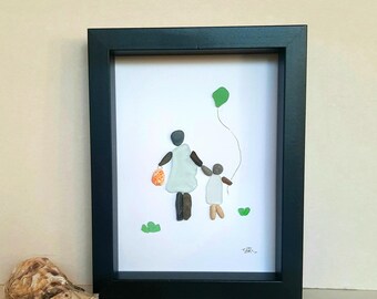 May include: Framed pebble art depicting a parent and child holding hands, with a green balloon. The figures are made of pebbles, wearing white dresses, and walking on a white background. The frame is black.