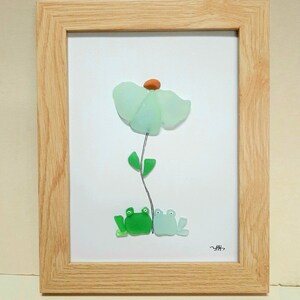 Peut inclure: Une œuvre d'art encadrée représentant une fleur en verre bleu clair avec une tige et des feuilles vertes. Deux grenouilles en verre vert sont assises à la base de la fleur.