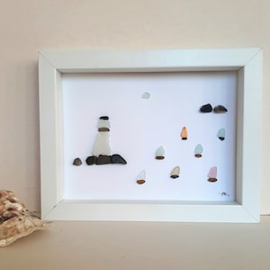 Peut inclure: Une image encadrée en blanc avec un phare et des voiliers en verre de mer et en pierres. L'image est sur un fond beige clair.