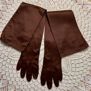 May include: A pair of long, dark brown gloves with a matching scarf. The gloves have a smooth texture and extend past the wrist. The scarf is long and rectangular, with a similar color and material as the gloves. The items are displayed on a white lace surface.