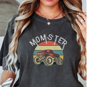 以下が含まれることがあります： 「MOMSTER」の文字と赤いモンスタートラックのグラフィックがプリントされた濃いグレーのTシャツ。トラックは、オレンジ、黄色、緑、青のストライプが入ったレトロな夕日のデザインを背景にしています。