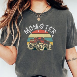 以下が含まれることがあります： 「MOMSTER」の文字と赤いモンスターカーのグラフィックがプリントされたダークグレーのTシャツ。グラフィックは、赤、黄、青の夕焼けを背景にしたレトロなデザインです。シャツは柔らかい素材で作られています。
