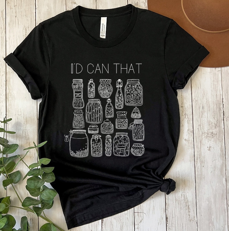 May include: Black t-shirt with white text that reads "I'd Can That" and a graphic of various jars with lids. The jars are filled with different items, such as pickles, beans, and herbs.