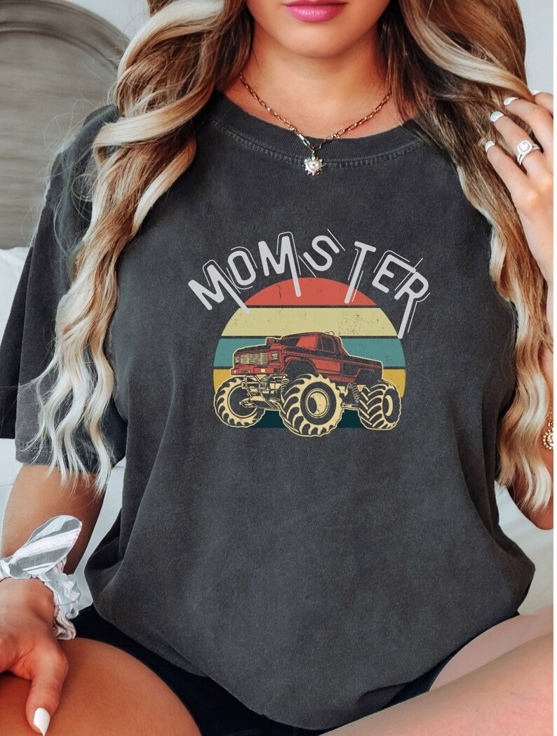 以下が含まれることがあります： 「MOMSTER」の文字と赤いモンスタートラックのグラフィックがプリントされた濃いグレーのTシャツ。トラックは、オレンジ、黄色、緑、青のストライプが入ったレトロな夕日のデザインを背景にしています。