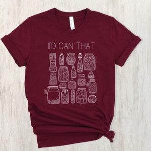 May include: A maroon t-shirt with the text "I'd Can That" in white lettering. The shirt features a design of various jars with different contents, all in white.