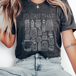 May include: A dark gray t-shirt with the text "I'd Can That" printed in white. The shirt features a design of various jars and bottles with white outlines.
