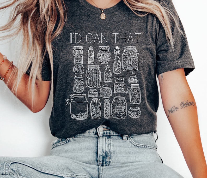 May include: A dark gray t-shirt with the text "I'd Can That" printed in white. The shirt features a design of various jars and bottles with white outlines.