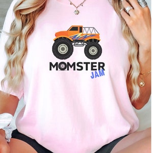 May include: A pink t-shirt with a graphic of an orange monster truck with blue and purple accents. The text "MOMSTER JAM" is printed below the truck.