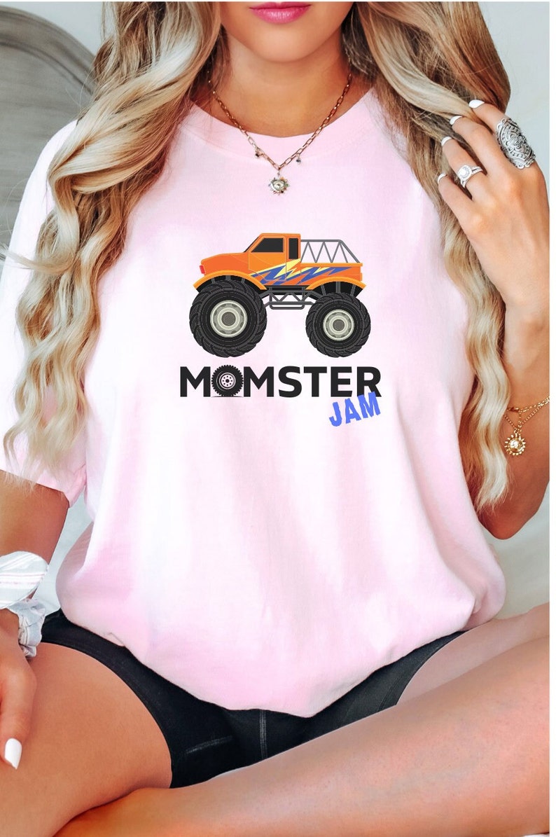 May include: A pink t-shirt with a graphic of an orange monster truck with blue and purple accents. The text "MOMSTER JAM" is printed below the truck.