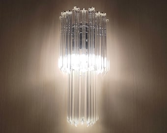 Peut inclure: Une applique murale en verre transparent au design moderne. L'applique est composée de plusieurs panneaux de verre verticaux qui créent un motif géométrique. La lumière est allumée et éclaire la pièce.