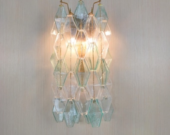 Peut inclure: Applique murale avec un design géométrique. Le luminaire est composé de pièces de verre en forme de losange transparentes et bleu clair, disposées verticalement. Il possède un cadre en métal couleur laiton et émet une lumière chaude.