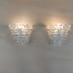 May include: A pair of clear, textured glass wall sconces with a tiered, faceted design. Each sconce features a glowing light source within the centre, held by small gold-toned metal clips on the sides, creating a bright, reflective illumination.