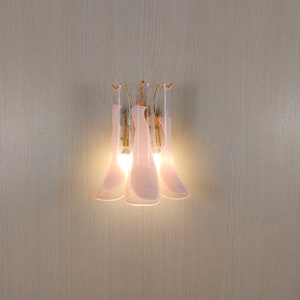 May include: A wall-mounted light fitting with three pink glass shades. The shades are shaped like petals and have a frosted finish. The light fitting is illuminated, casting a warm glow.