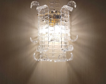 May include: A clear glass wall sconce with a geometric design. The sconce has a gold metal base and is lit by a light bulb.