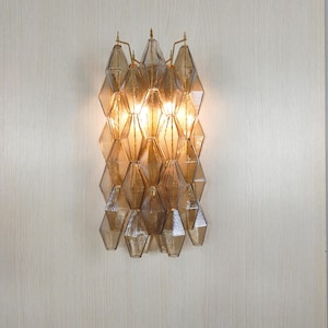 Puede incluir: Aplique de pared decorativo con diseño geométrico. Presenta múltiples paneles de vidrio en forma de diamante en tonos ámbar y marrón, dispuestos verticalmente. La fuente de luz es visible, creando un brillo cálido.