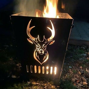 Op de afbeelding: Een zwarte metalen vuurkorf met een silhouet van een hertenkop uitgesneden op het voorpaneel. De vuurkorf is gevuld met brandend hout en vlammen zijn zichtbaar. De vuurkorf staat buiten, met een donkere achtergrond.
