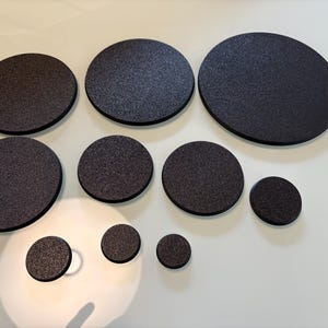 Könnte beinhalten: Eine Sammlung dunkelgrauer, runder Untersetzer in verschiedenen Größen. Die Untersetzer haben eine strukturierte Oberfläche und ein schlichtes, modernes Design. Sie sind auf einer weißen Oberfläche angeordnet und zeigen ihre unterschiedlichen Durchmesser.