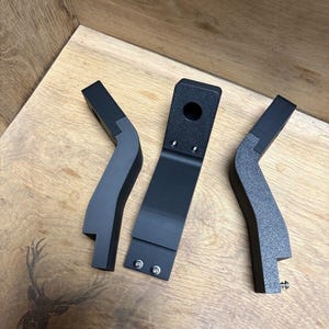 Könnte beinhalten: Drei schwarze Kunststoffhalterungen mit einer strukturierten Oberfläche. Die Halterungen sind abgewinkelt und haben ein Loch in der Mitte der Oberseite.