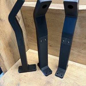 Könnte beinhalten: Drei schwarze Plastik-Tischbeine mit einem geschwungenen Design und einem Loch oben. Die Beine stehen auf einer Holzoberfläche.