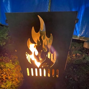Könnte beinhalten: Ein schwarzer Metall-Feuerkorb mit einem flammenförmigen Ausschnitt. Der Feuerkorb hat eine rechteckige Form mit vertikalen Schlitzen am Boden. Helle Flammen sind im Inneren sichtbar und werfen einen warmen Schein.