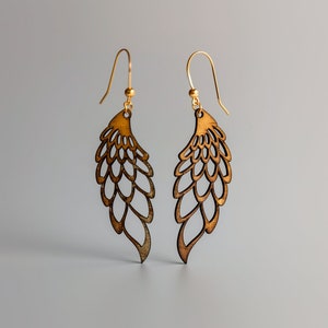 Puede incluir: Un par de pendientes de oro con un diseño de madera marrón. Los pendientes tienen forma de alas con un patrón delicado e intrincado.