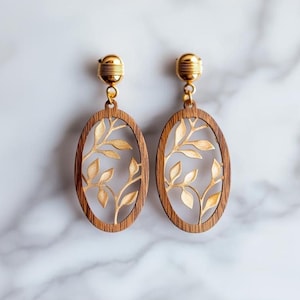 May include: A pair of oval-shaped wooden earrings with a gold leaf design. The earrings are made of dark wood with a gold-colored metal accent.