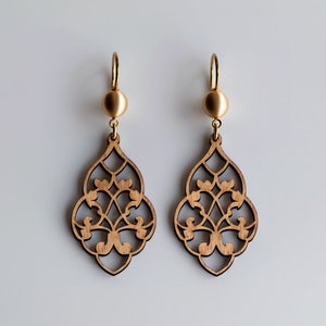 以下が含まれることがあります： 繊細で複雑なデザインの金と木のイヤリングのペア。イヤリングは、上部に金色のビーズ、下に花柄のウッドペンダントが付いています。