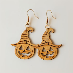 May include: A pair of wooden earrings shaped like jack-o'-lanterns wearing witch hats. The earrings are a light brown color and have gold hooks.