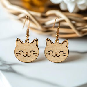 May include: A pair of gold cat-shaped earrings with a smiling face. The earrings are made of wood and have a light brown color.
