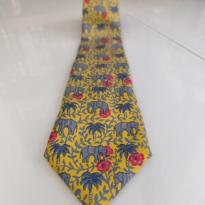 Pode incluir: Uma gravata amarela com um padrão repetitivo de elefantes cinzentos, palmeiras azuis e pequenas flores vermelhas. O design é apresentado sobre um fundo de finas vinhas azuis que ligam os vários elementos por toda a superfície do tecido.
