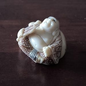 Peut inclure: Figurine ivoire d'un Bouddha souriant assis, tenant un éventail. La robe du Bouddha présente un motif texturé et détaillé. La figurine est sur une surface marron foncé.