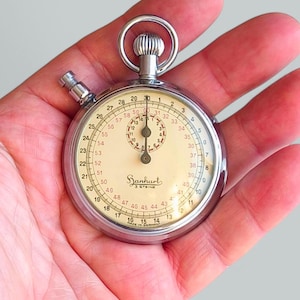 Puede incluir: Un cronómetro de bolsillo plateado con una esfera blanca y marcas negras. El reloj tiene una aguja negra y una aguja de segundos roja. El reloj está hecho en Alemania y tiene el nombre de la marca "Sanhart 3 Steine" impreso en la esfera.