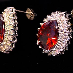 Op de afbeelding: Een paar oorstekers met grote, peervormige, dieprode edelstenen. Elke oorbel is omgeven door een halo van kleinere, heldere edelstenen gezet in een zilverkleurig metaal. De oorbellen hebben een stift en vlindersluiting.