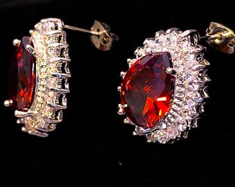 May include: A pair of stud earrings featuring large, pear-shaped, deep red gemstones. Each earring is surrounded by a halo of smaller, clear gemstones set in a silver-colored metal. The earrings have a post and butterfly backing.