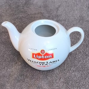 Puede incluir: Tetera de cerámica blanca con un logotipo de té Lipton Yellow Label rojo y negro. La tetera tiene un asa y un pico.
